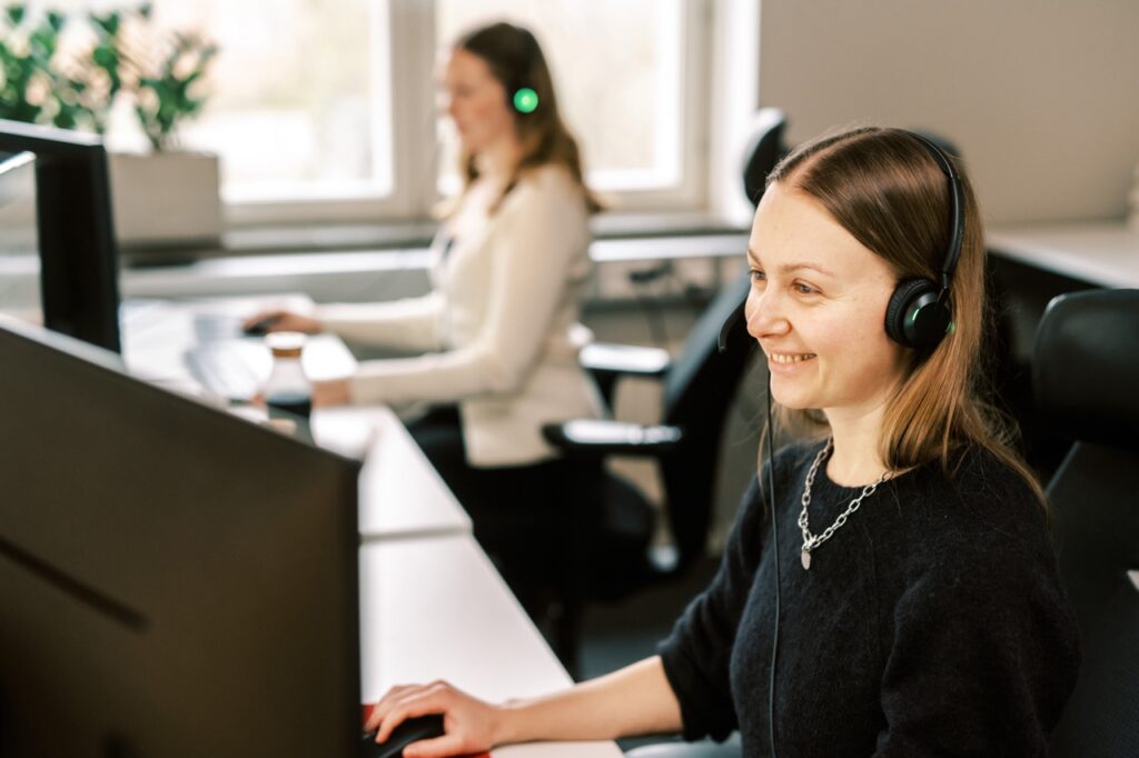 Kvinna på beställningscentral sitter framför datorn leendes och med ett headset på huvudet.